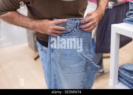 Person in a casual brown shirt comparing the waist of a light blue denim jean with their current size, indicating careful consideration during clothes Stock Photo