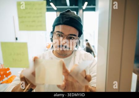 Male employee sticking adhesive note on glass wall inside meeting room Stock Photo