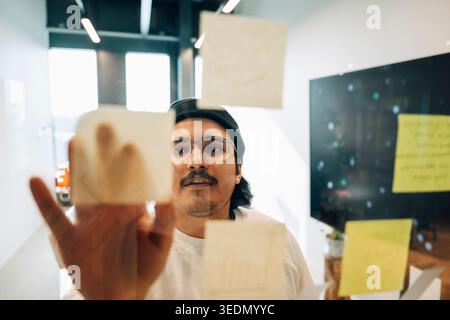 Young male business professional sticking adhesive notes on glass wall at office Stock Photo