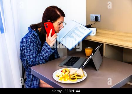 Young woman multitasking with book, phone, and laptop while having breakfast Stock Photo