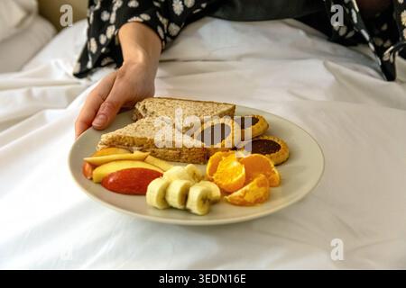 Person holding breakfast plate with sandwiches and fruit in bed Stock Photo
