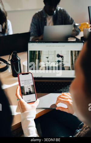 Over the shoulder view of businesswoman comparing websites on phone and laptop while working at office Stock Photo