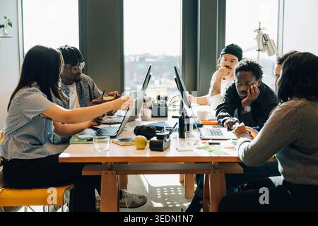 Diverse team of programmers working on laptops and computers while sitting around cluttered desk in office Stock Photo