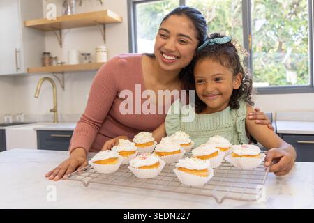 Mother and daughter in pink top, green dress smiling presenting cupcakes on cooling rack in kitchen Stock Photo
