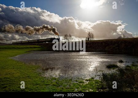 A steam train on a tall embankment of a heritage railway with flooded field in the foreground and bight sunlit sky, East Lancashire Railway, Bury. Stock Photo