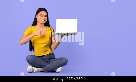 Smiling Student Shows Laptop While Sitting on Floor in Studio With Purple Background Stock Photo