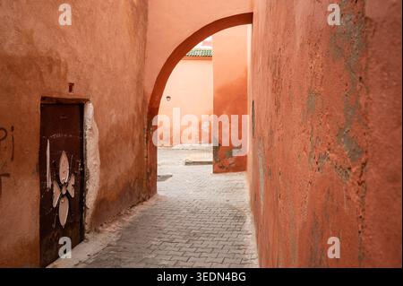 Winding back alley in the Marrakech medina framed by an arched passage, orange textured walls and a weathered wooden door. Atmosphere feels historic a Stock Photo