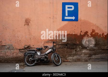 An old motorcycle leans against a weathered terracotta wall beneath a blue parking sign in a narrow Marrakesh street, evoking urban decay, travel atmo Stock Photo