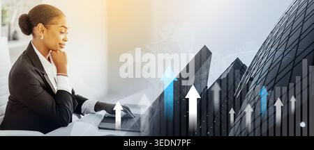 Confident black businesswoman looking at laptop and smiling, sitting at workplace in office, side view, free space Stock Photo