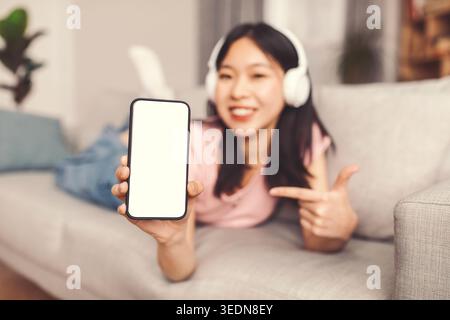 Young Woman Points at Smartphone Screen While Lying on Couch in Living Room Stock Photo