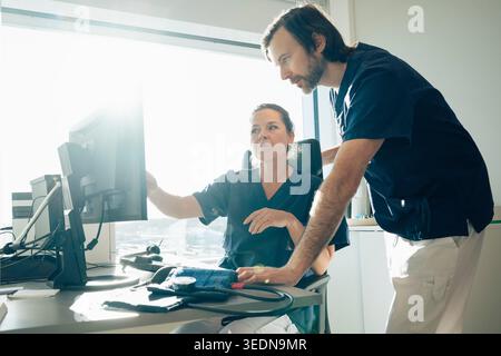Female doctor discussing medical record with male coworker on computer while sitting in office at hospital Stock Photo