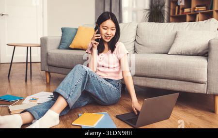 Young Woman Talks on Phone While Sitting on Floor and Using Laptop in Living Room Stock Photo