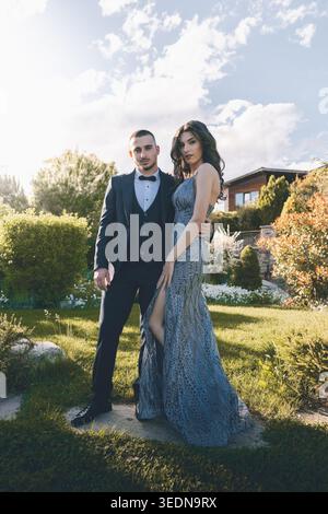 Ognyanovo, Bulgaria - 06 24 2025: Stylish young couple dressed in formal evening attire posing together in a landscaped garden during golden hour. Rom Stock Photo