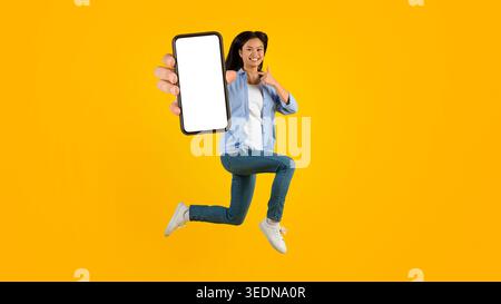 Woman Jumps and Shows Smartphone in Bright Studio With Yellow Background Stock Photo