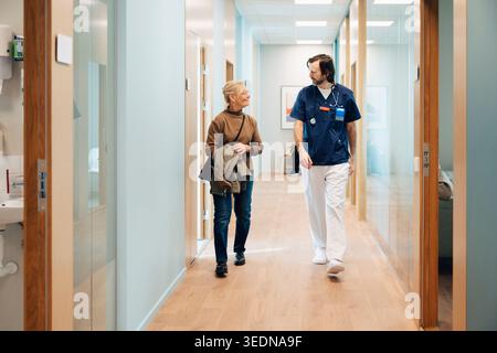 Full length of male medical professional talking with senior woman while walking in hospital lobby Stock Photo