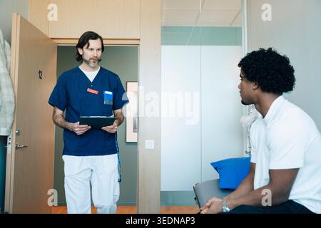 Male doctor wearing scrubs walking towards young patient sitting on table in examination room at clinic Stock Photo