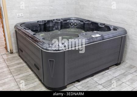 Modern dark grey hot tub filled with water in a luxury indoor spa center. The square jacuzzi is surrounded by light marble tiles, creating a relaxing Stock Photo