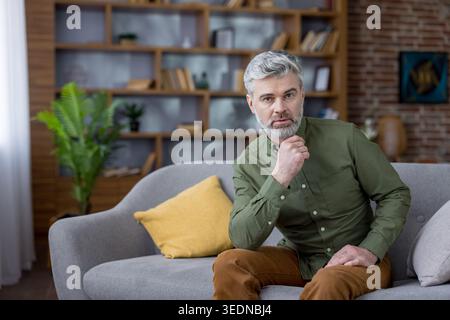 Mature man with gray hair and beard sitting on a comfortable gray sofa, looking confidently at the camera while thoughtfully resting his hand on his chin in a stylish home interior Stock Photo