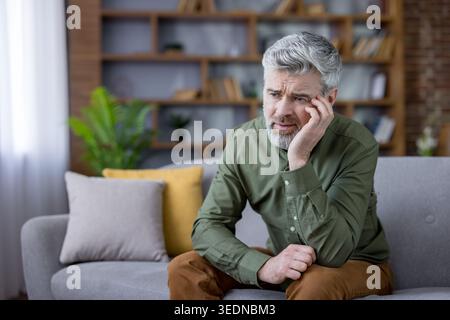 Senior man feeling upset and worried while sitting on a comfortable sofa at home, experiencing feelings of loneliness and mental health struggles related to aging Stock Photo