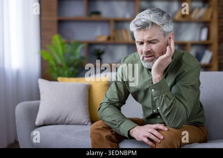 Mature man with gray beard sitting on a couch, holding his ear in discomfort, grimacing from ear pain or tinnitus in a modern living room, appearing worried and tired Stock Photo