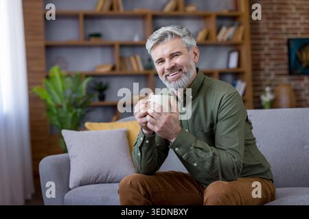 Mature man with a silver beard smiling at the camera, relaxing on a gray sofa in a modern living room, a bookshelf visible in the background, having a hot drink Stock Photo