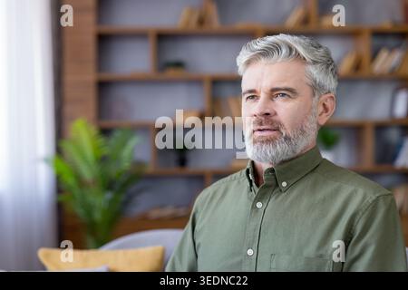 Mature grey-haired man with beard in casual shirt, gazing thoughtfully away in a modern home office with bookshelf and plants, relaxed and focused during remote work Stock Photo