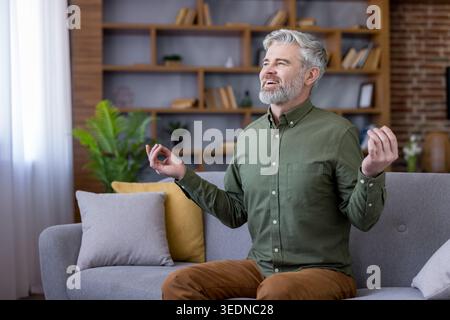 Senior man with gray hair and beard is meditating in a calm living room, practicing mindfulness and finding peace for mental wellbeing and stress relief Stock Photo