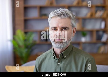 Confident middle-aged man with grey hair and beard sitting in living room or home office against blurred background, showing professionalism, experience, and remote work Stock Photo