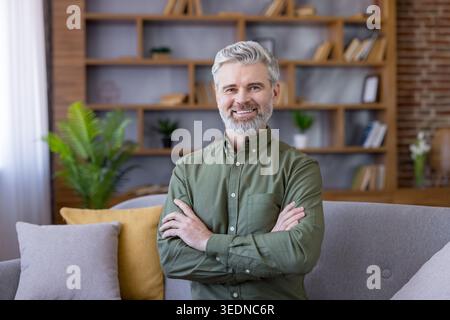 Mature man with gray hair and beard smiling, standing with crossed arms in a cozy living room, symbolizing confidence, success, and a comfortable lifestyle Stock Photo