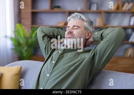 Mature man with gray hair and beard relaxing on a cozy sofa at home, eyes closed, hands behind head, enjoying a peaceful, mindful moment of calm and contentment Stock Photo