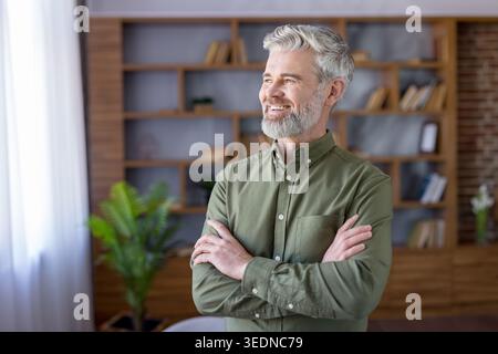 Confident senior man wearing a green shirt, with gray hair and a beard, standing proudly in a modern home office setting while looking thoughtfully into the distance Stock Photo
