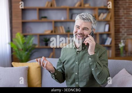Happy mature man using a smartphone for a conversation, expressing joy and confidence while sitting comfortably at home and engaging in modern communication Stock Photo