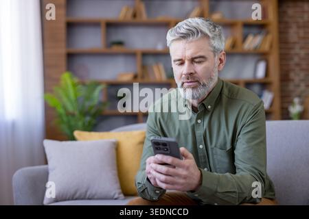 Mature man with grey hair and beard sitting on a sofa, browsing content on a mobile phone in a modern living room while enjoying digital communication and entertainment Stock Photo