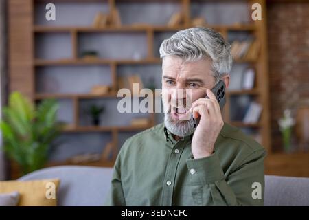 Mature man with grey hair and beard looking angry and yelling into his smartphone while having a stressful conversation, sitting on a couch in a home or office setting Stock Photo