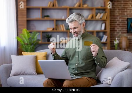 Mature bearded man with gray hair celebrates online shopping success on his laptop at home, holding a gold credit card with a joyful, satisfied expression on the sofa Stock Photo