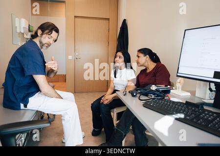Male pediatrician explaining condition to mother and daughter while sitting on examination table in clinic Stock Photo