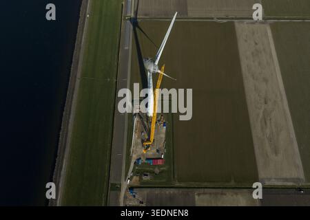 Aerial view of a large white wind turbine being serviced by a tall yellow crane in a flat agricultural field next to a canal with red containers at th Stock Photo