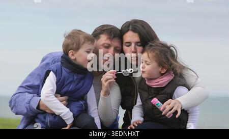 Parents and children enjoying playful family time together with soap bubbles outdoors in a park on a cloudy autumn day. Stock Photo
