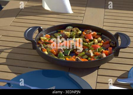 A rustic presentation of grilled peppers, mushrooms, and zucchini served in a cast iron pan on a sunny wooden patio table. Stock Photo