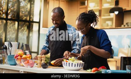 African american man seasoning the salad with mix of spices and pepper, pouring condiments for a tasty vegetarian meal. Smiling woman mixing fresh vegetables together, healthy lifestyle. Stock Photo
