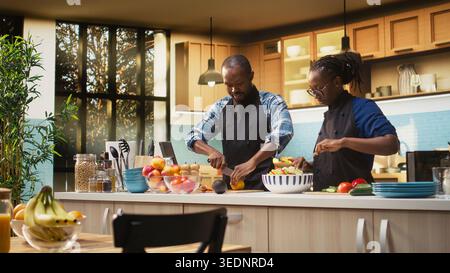 Happy black man squeezing lemon juice over fresh vegetables, preparing a tasty salad together with girlfriend. Seasoning lunch meal with bio organic ingredients, relaxed home moment. Stock Photo