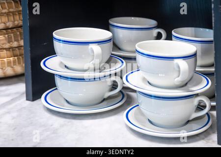 Stacks of white ceramic coffee cups with blue stripes and saucers on a shelf in a shop. Stock Photo