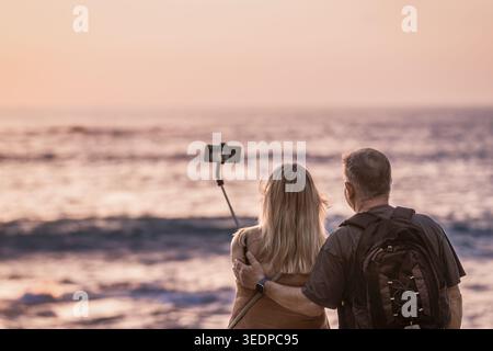 Mature couple taking selfie on the beach at sunset with smartphone and selfie stick, standing together near the sea while looking at the ocean. Warm e Stock Photo
