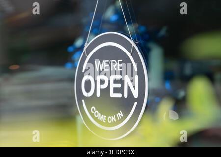 Round We are open sign hanging on glass storefront or door indicating business is open and ready to welcome customers. Blurred blue and green backgrou Stock Photo