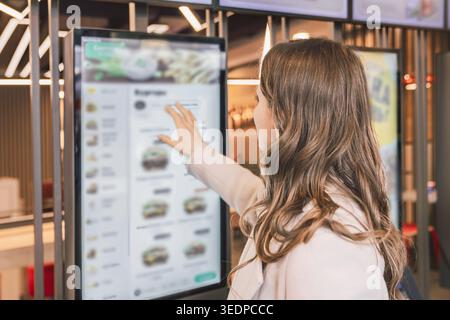 A woman interacts with a large touchscreen kiosk in a modern fast food restaurant, selecting items from a digital menu. The scene reflects the growing Stock Photo