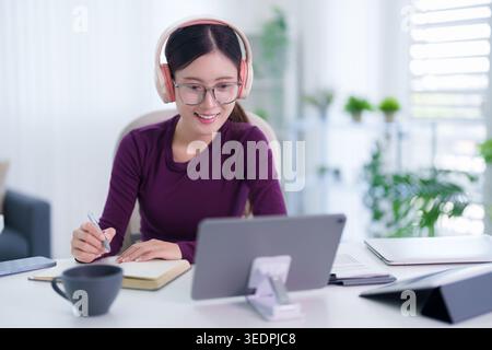 Young Asian female student wearing headphones and glasses using tablet computer taking notes at home office workspace Stock Photo