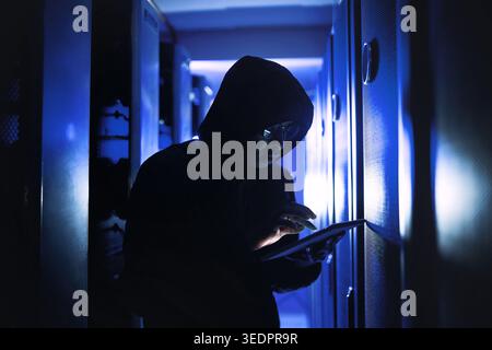 Person, hacker or night with tablet in server room for malware attack, network breach or crime. Criminal, cracker or cyberpunk with technology for Stock Photo