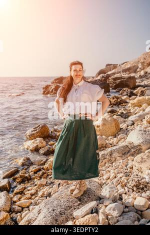 Woman coastline sunset smiling lady in white blouse and green skirt stands on rocky beach during golden hour travel mood Stock Photo