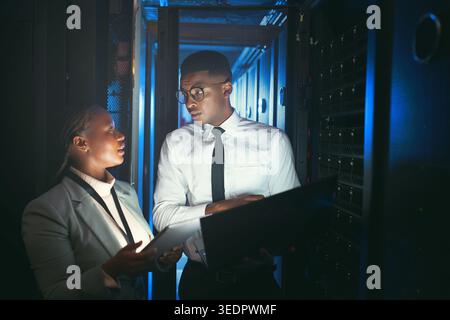 Inspection, review or technicians in server room with tech, teamwork or system check in data center. Engineer, laptop or black people in dark Stock Photo