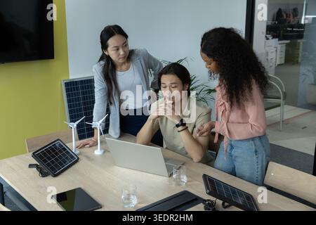 Diverse coworkers in business attire collaborating at office table using laptop and solar panels Stock Photo